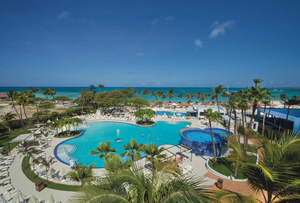 A tropical resort pool area with a large blue pool, surrounding lounge chairs, palm trees, and a beach and turquoise ocean in the distance.
