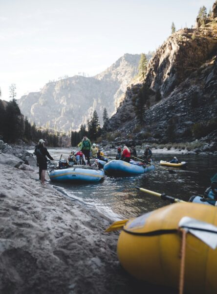 Group of people preparing inflatable rafts on a rocky riverbank in a mountainous canyon with pine trees and steep cliffs in the background.
