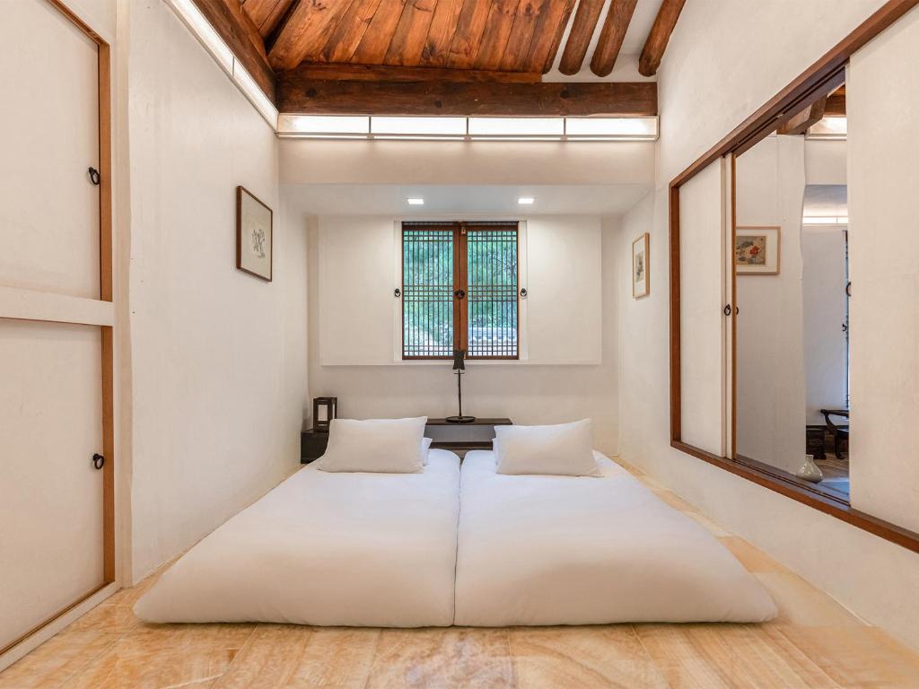 Minimalist bedroom with a low white bed on a warm tile floor and two pillows under a wooden-beam ceiling.