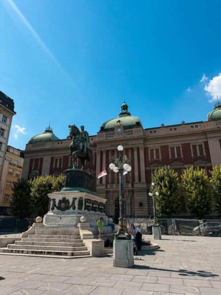Equestrian statue in front of a grand neoclassical museum building with green domes under a bright blue sky in a city square.