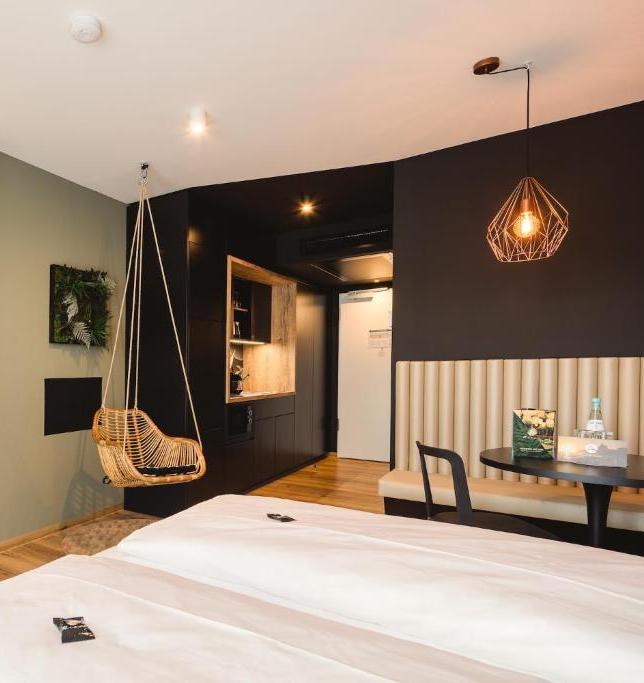 Hotel room scene with white beds in the foreground, a hanging wicker chair, dark kitchen wall, round table and bench seating under a geometric pendant light.