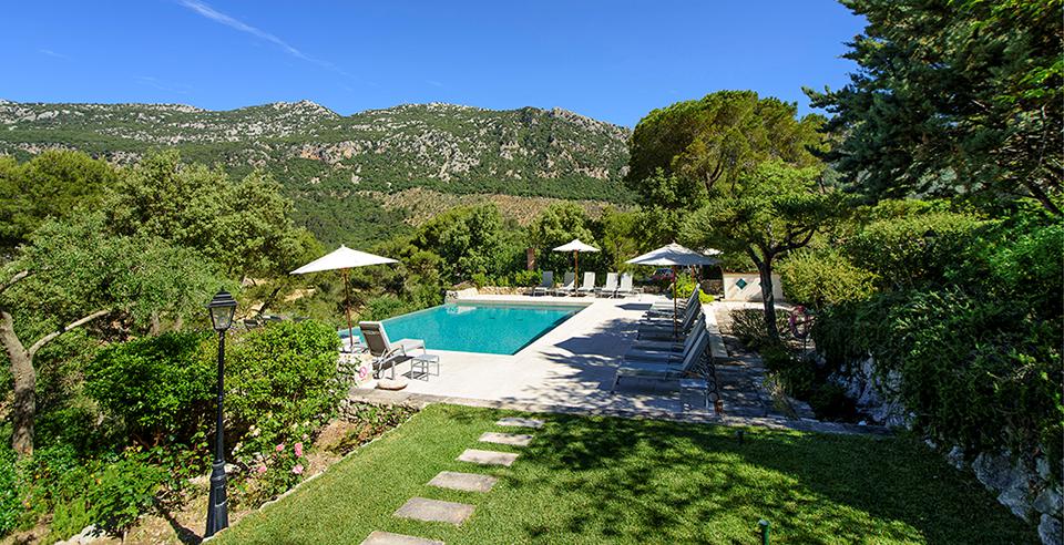 Outdoor pool area with white umbrellas and lounge chairs, set in a garden with hills in the background.