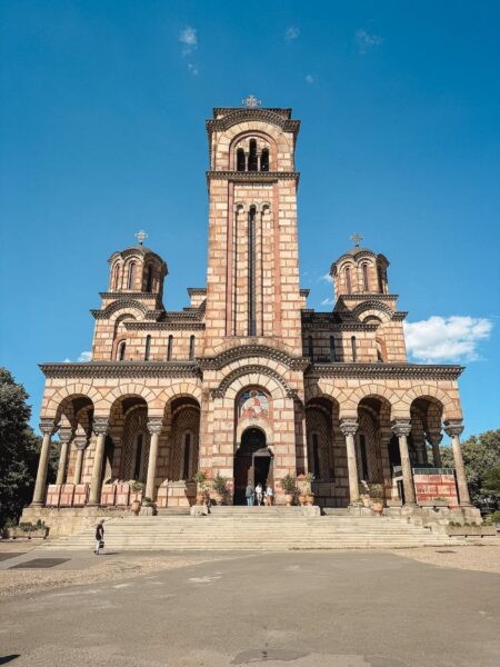 Facade of a brick church with arches and tall central bell tower under a clear blue sky, with two smaller domed towers on each side and steps leading up to the entrance