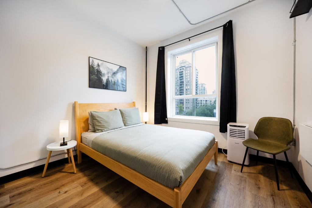 Cozy bedroom with a wooden bed, gray bedding, and two pillows beside a small round nightstand lamp. A large window with black curtains overlooks city buildings.