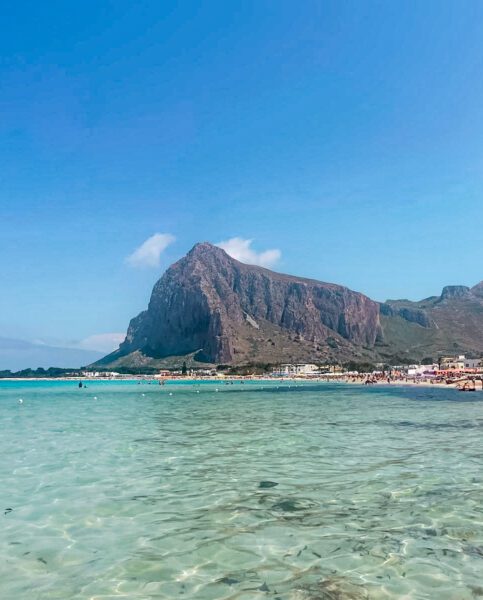 Clear turquoise sea at a sunny beach with a towering rocky mountain in the background and people along the shore.