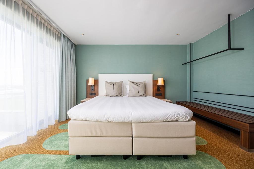 Hotel bedroom with a large white-duvet bed, beige upholstered base, and two bedside lamps on wooden nightstands against a teal wall