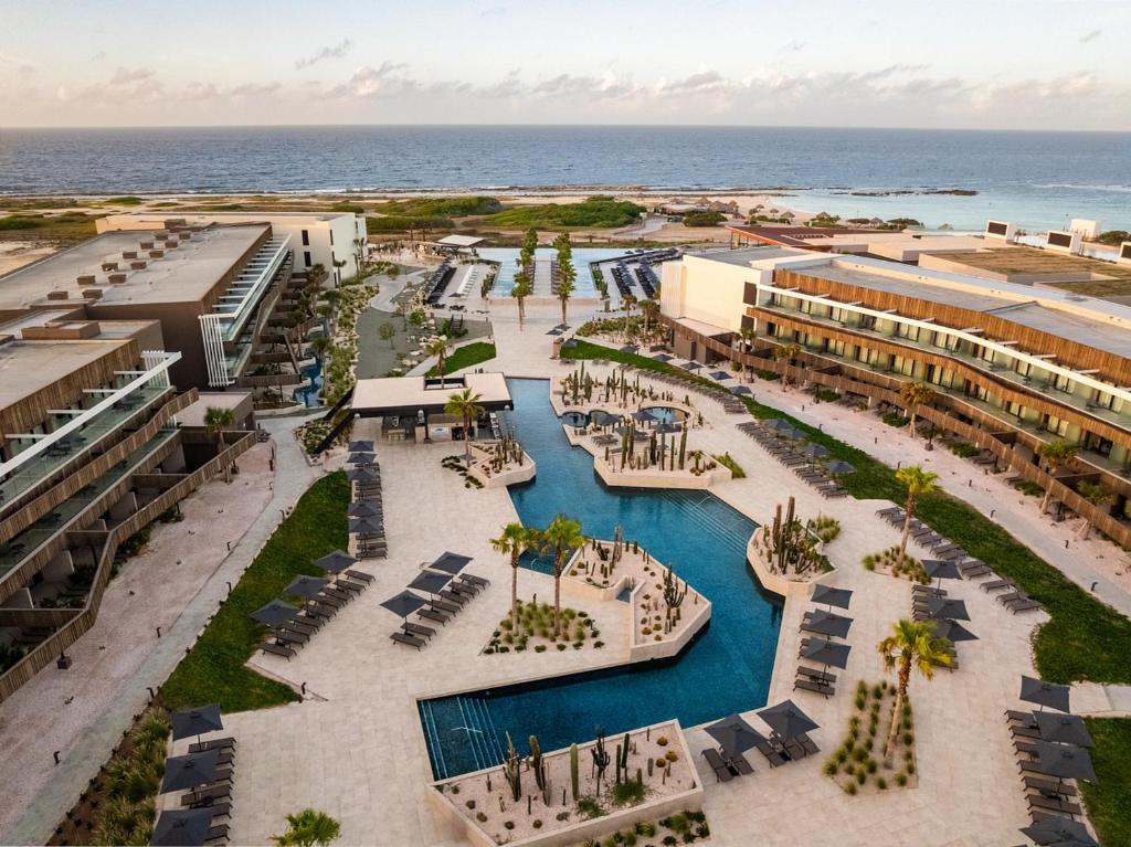 Aerial view of a resort with multiple pools, palm trees, and lounge chairs surrounding geometric pools, ocean in the distance.