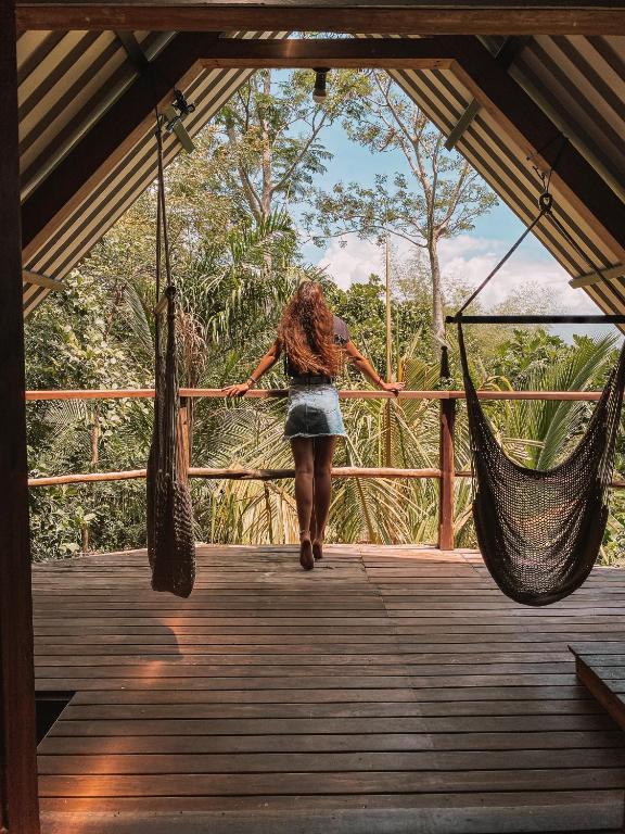 Person with long hair wearing a denim skirt standing with their back to the camera, leaning on a wooden railing of a treehouse deck and looking into a tropical jungle. Two black hammocks hang from ropes on either side of the deck.