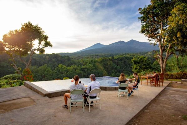 Group of people seated on folding chairs around an infinity pool, with a forest and distant mountains at sunset in the background.