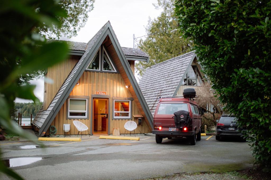 A-frame wooden office building with a triangular roof, two white chairs outside, and a red van parked nearby in a parking lot.