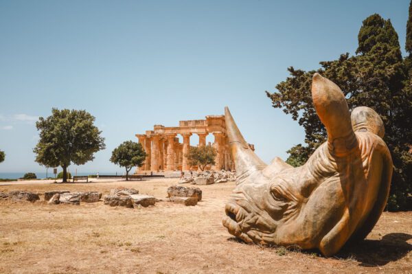 Ancient temple ruin with tall columns by the sea, foreground dominated by a large stone sculpture resembling a sea creature's head under a clear blue sky.