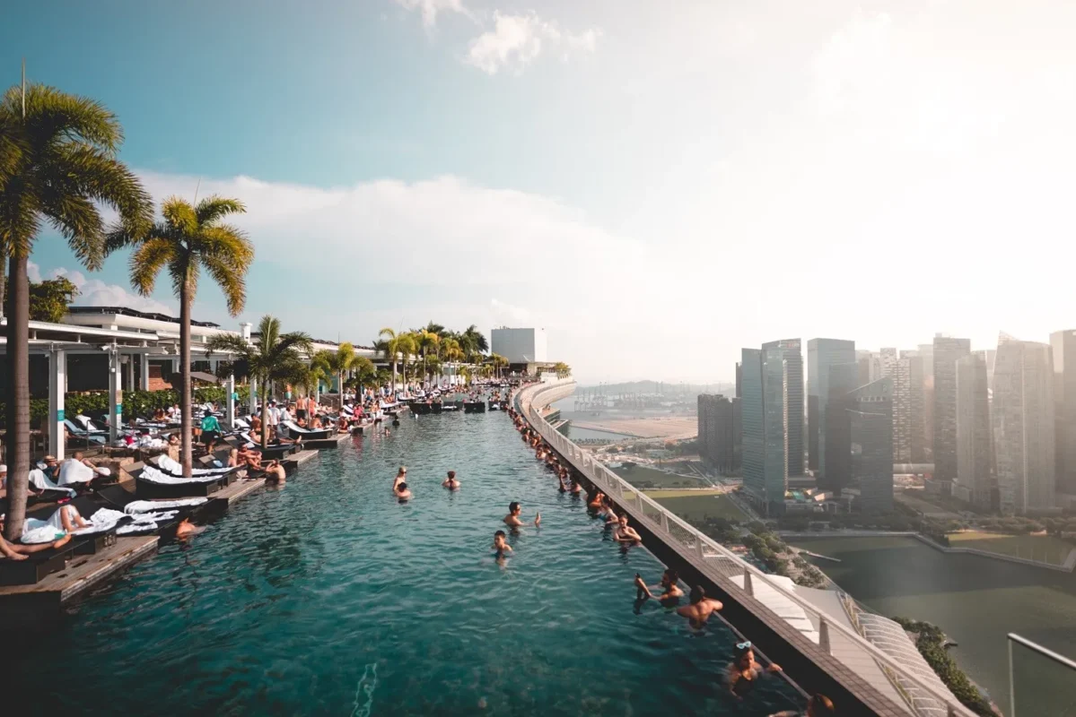 People lounging and swimming at an outdoor rooftop infinity pool with palm trees and a city skyline.