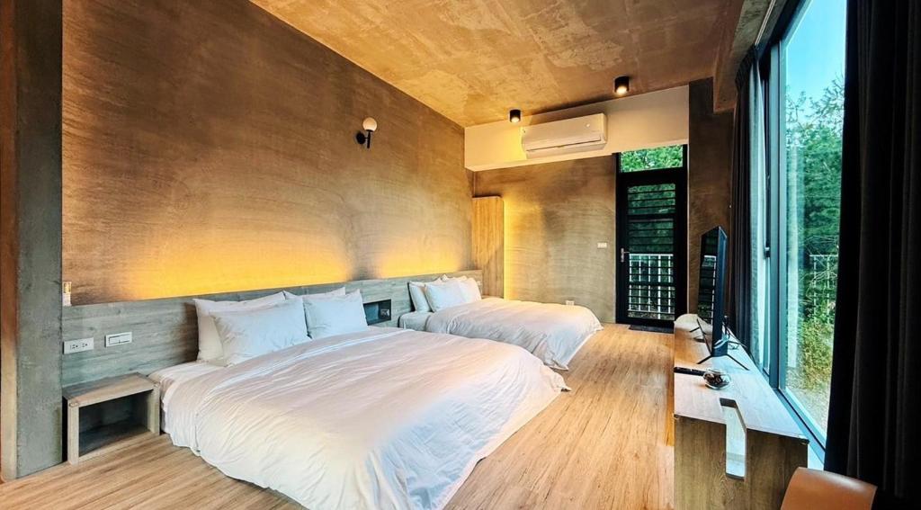 Two white beds in a modern concrete-walled hotel room with a wooden floor and a large window overlooking greenery, TV on a low console nearby, and an edge-lit wall light.