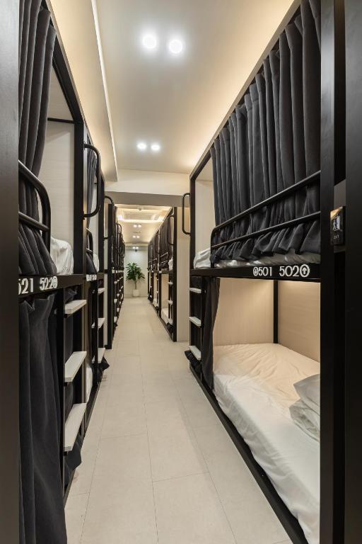 Long dormitory hall with two-tier black metal bunk beds along both sides, numbered beds, white linens, and a plant at the far end.
