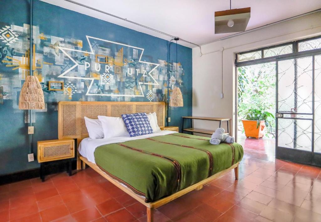 Bedroom with a wooden bed and green bedspread, woven headboard, blue geometric wall art, wicker pendant lights, and a glass door opening to a sunny patio with potted plants.