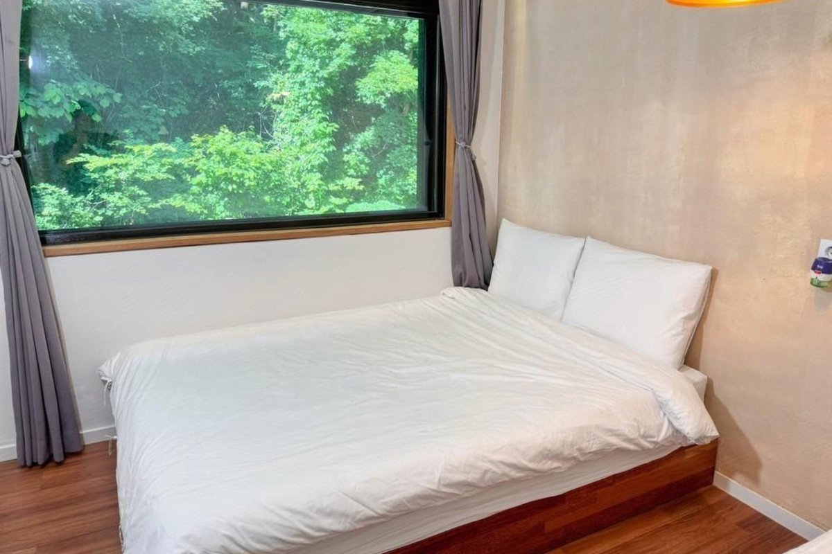 Modern bedroom with a large white bed by a window overlooking green trees, framed by grey curtains.