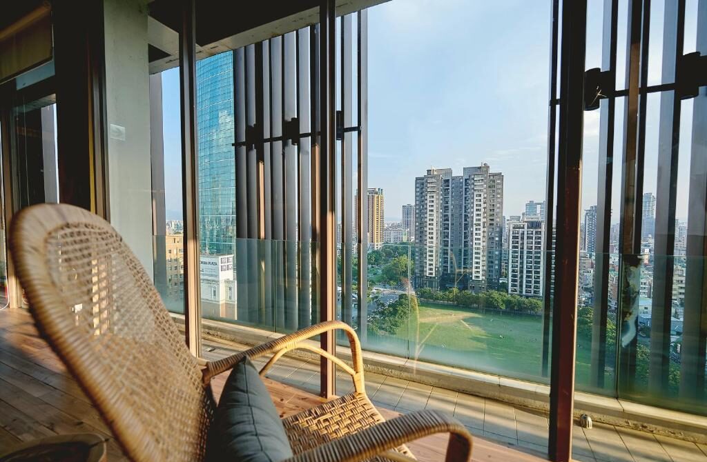 Wicker lounge chair on a balcony overlooking Taichung's city skyline and a green park beneath a clear blue sky.