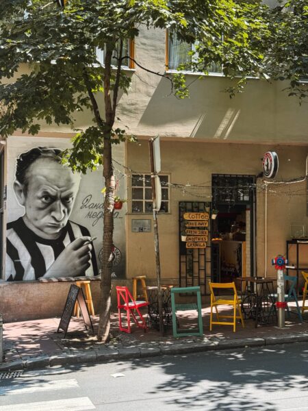 Cafe storefront with colorful outdoor chairs and a large black‑and‑white portrait on the left wall under a tree, street scene.