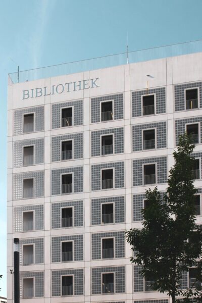 Facade of a modern library building with the word BIBLIOTHEK on the top, rows of square windows with glass blocks, and a tree in the foreground.