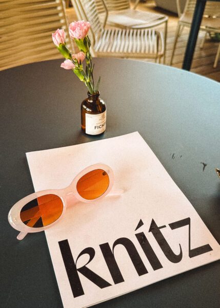 Amber glass bottle with pink carnations on a dark round table, with pink sunglasses and a magazine nearby.