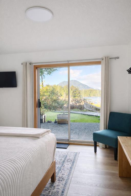 Bedroom with a sliding glass door that opens to a grassy yard and lake with mountains in the distance; a bed on the left and a teal chair with a wooden desk on the right.
