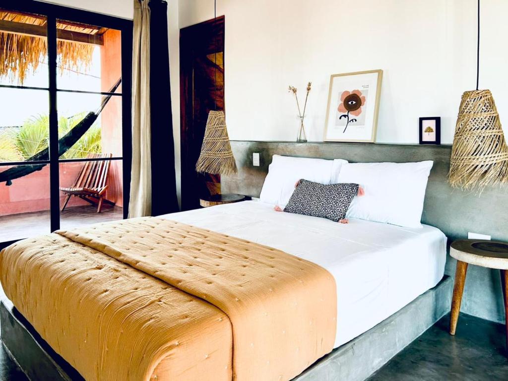 A modern hotel room with a made bed, white linens, an orange quilt, and woven pendant lamps, opens to a balcony with a hammock chair.