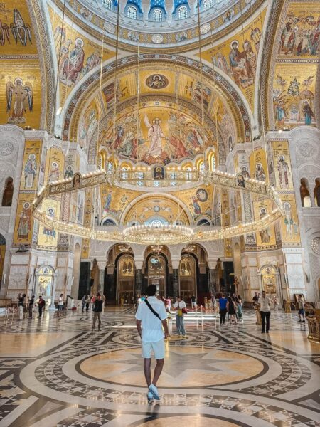 Interior of a grand Orthodox cathedral with gold mosaics on domed ceilings and arches, chandeliers, and visitors scattered on a marble floor.