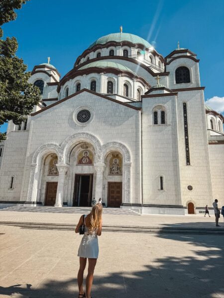 Wide-angle view of a white Orthodox church with green domes under a clear blue sky, a woman in a white dress stands in the foreground.