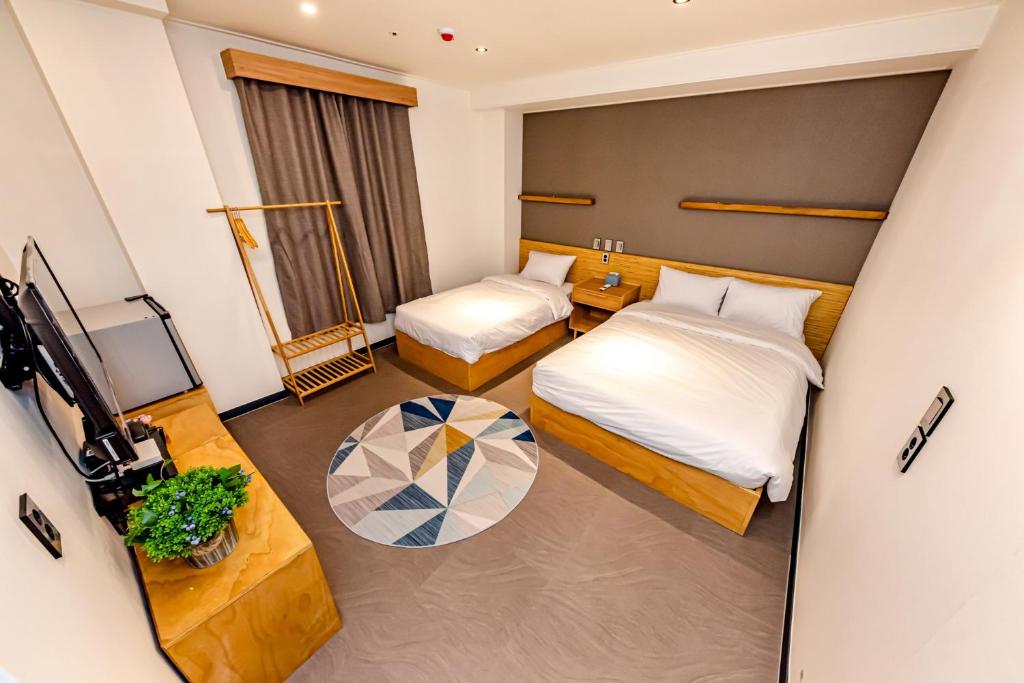 Hotel room with two twin beds and white linens, wooden headboards, a shared nightstand, and a geometric rug in the center. A clothing rack sits by a large window with dark curtains; a potted plant and a wall-mounted TV are visible opposite the beds.