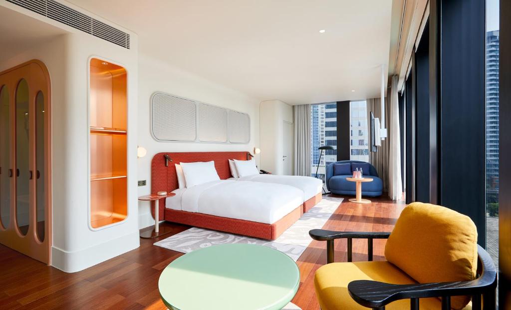 Contemporary hotel room with two white beds, orange headboard, and a seating area by large windows overlooking the city.