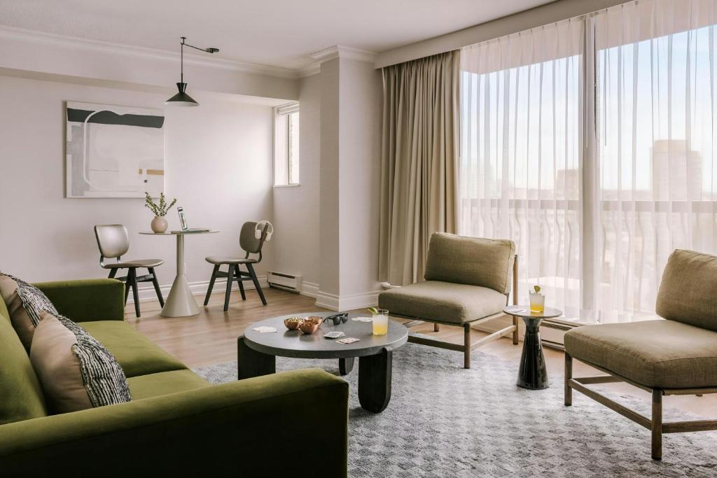 Modern living room with an olive-green sofa, beige lounge chairs, and a round coffee table; sheer-curtained balcony in the background.