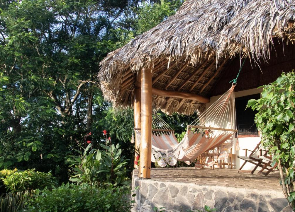 Hammock hanging between wooden posts on a thatched-roof porch surrounded by tropical plants and greenery