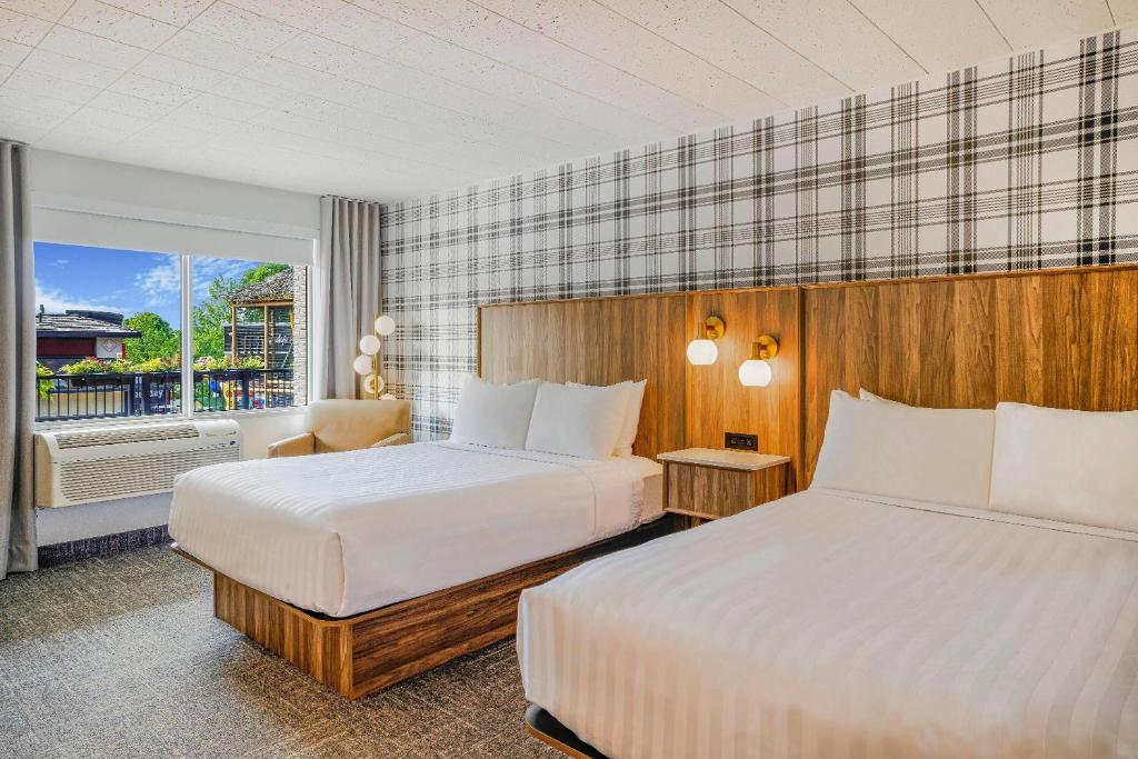 Hotel room with two white beds, wood headboard, plaid accent wall, and a window view behind a chair, sunlit.