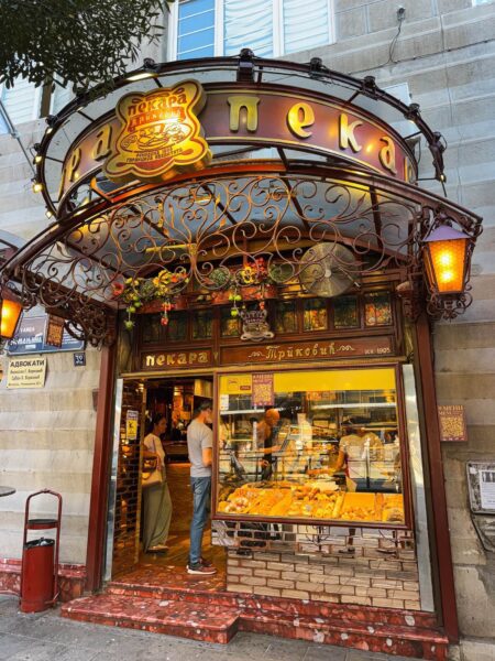 Bakery storefront with ornate wrought-iron arch, warm lights, and a display window of baked goods inside (Cyrillic signs above).