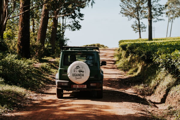 Rear view of a green SUV with a spare-tire cover reading 'Uganda Nomads' on a dirt rural road lined with trees and hedges