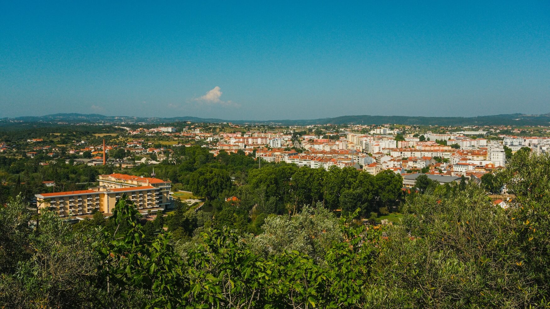 Panoramic view of a town with red-roofed buildings surrounded by trees and hills under a clear blue sky
