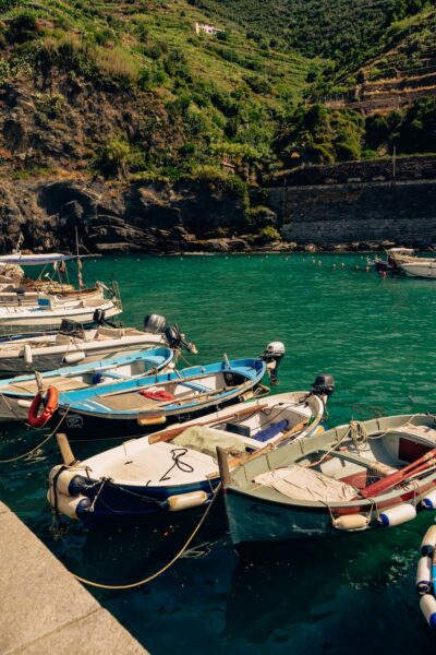 Several small boats moored along a turquoise harbor, with a green, terraced hillside and stone seawall in the background.