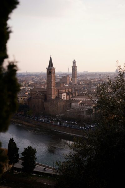 Aerial view of a historic Italian city with two tall brick towers along a river at sunset, framed by trees.