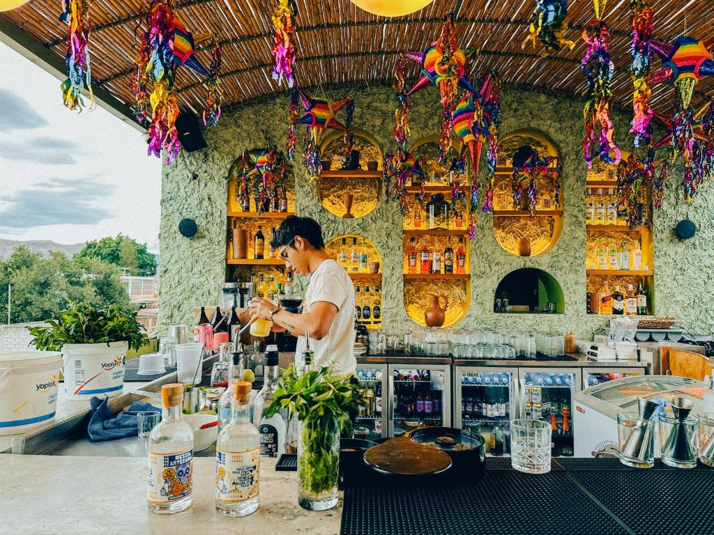 Bartender mixing a drink at an open-air bar with colorful hanging decorations and a lime-green textured wall.