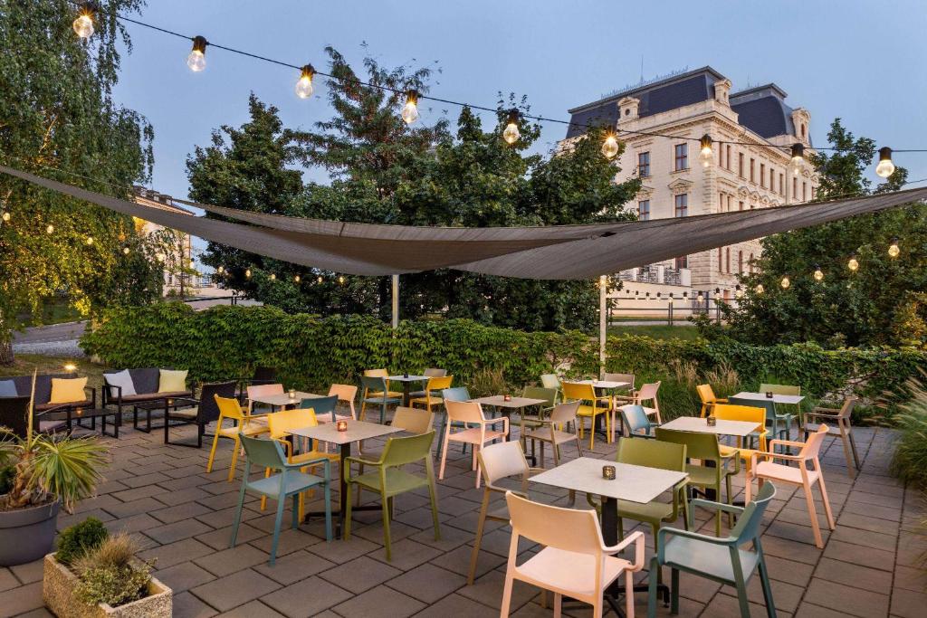 Outdoor cafe courtyard with white tables, colorful plastic chairs, string lights, and a shade sail in an urban setting at dusk.
