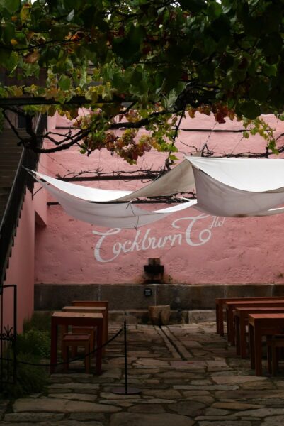 Pink courtyard with white sail-shaped canopies over wooden tables; stone path leads through seating and a pink wall with 'Cockburn Ltd' mural.