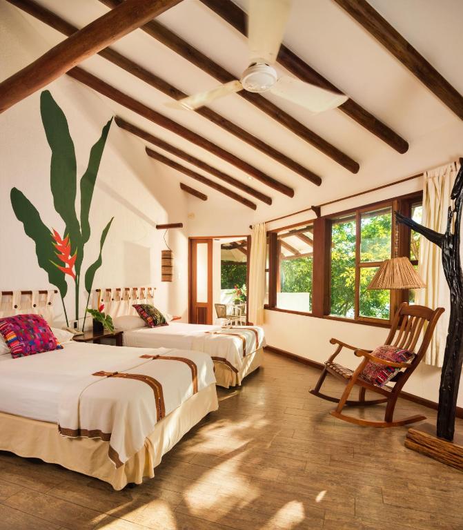 Hotel room with two twin beds, tropical leaf mural, ceiling fan, and a rocking chair by a large window.