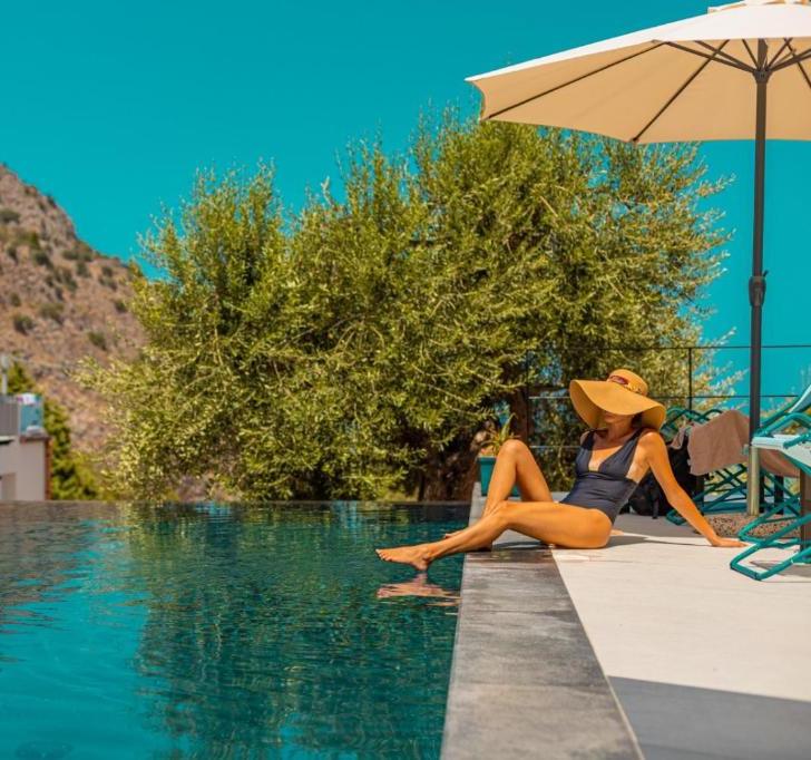 Woman in a black swimsuit sits on the pool’s edge with feet in the water under a large beige umbrella, relaxing by a turquoise pool.