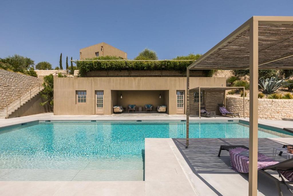 Modern backyard pool area with tan cabana, loungers, and a stone wall under a clear blue sky.