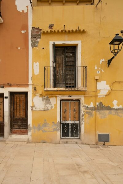 A yellow stucco building with peeling paint, featuring a small balcony with a black metal railing above a weathered door and a window with decorative grilles on a worn wall.