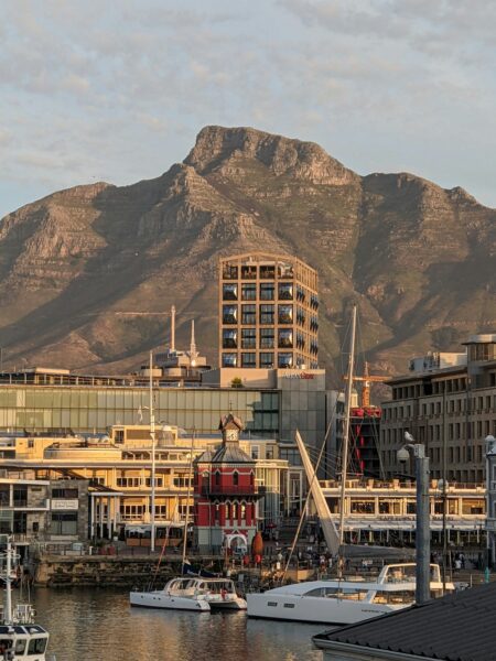 Harbor with moored yachts and sailboats, red brick tower, and glass office building against a rugged mountain backdrop at sunset sunlit.