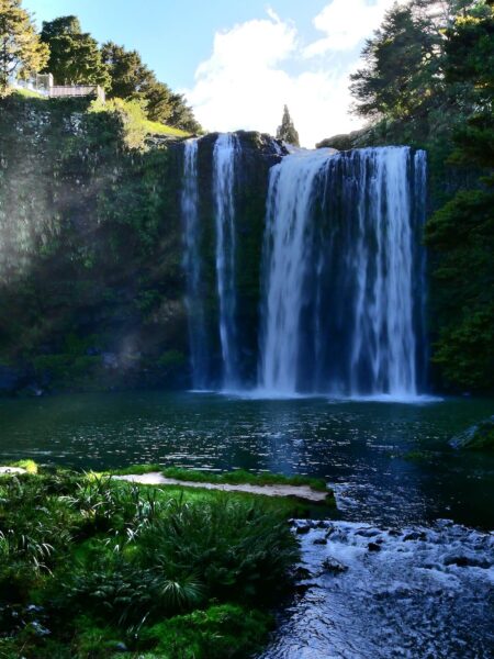 Waterfall tumbling over rocks into a tranquil pool, framed by lush green trees and a sunlit sky above.