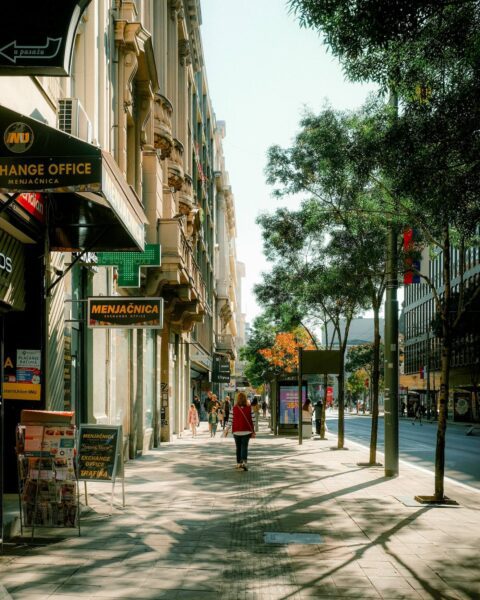 Urban street with shops on the left and trees lining the sidewalk; signs read 'EXCHANGE OFFICE' and 'MENJAČNICA'.