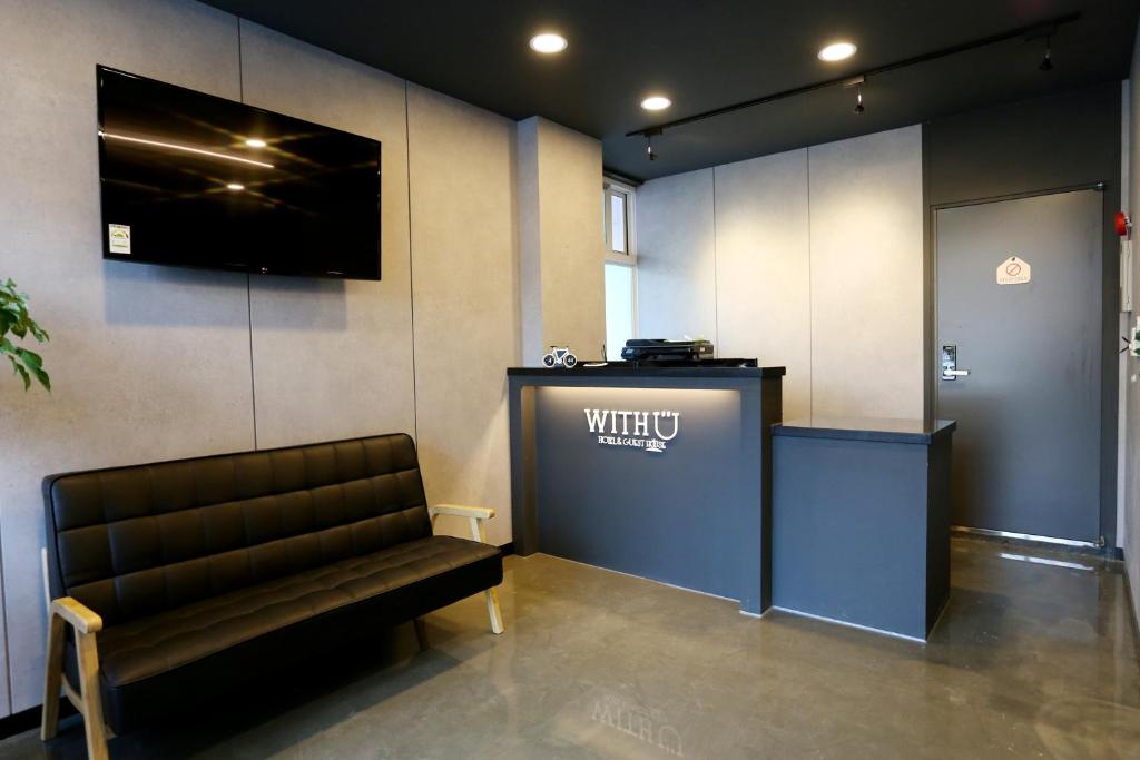 Hotel reception area with a blue front desk displaying the 'WITH U' logo, overhead lights, and a black leather bench in the lobby
