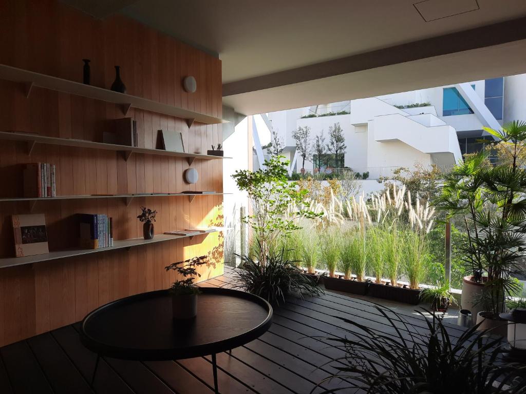 Interior balcony with wooden wall shelves, a round table, and potted plant, opening to a sunlit terrace with ornamental grasses and a white modern building beyond.